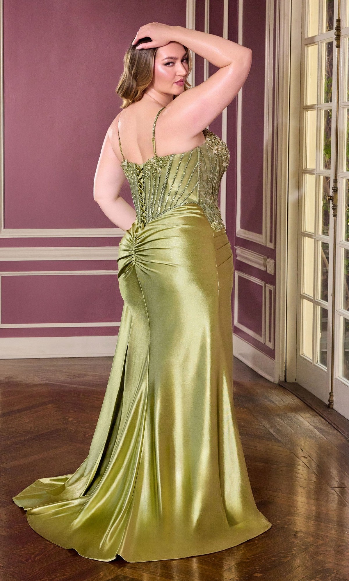 A woman wears the Ladivine CDS556C, a long plus-size fitted olive green satin prom dress with an embellished bodice and ruched back, standing in a room with mauve walls and white trim.